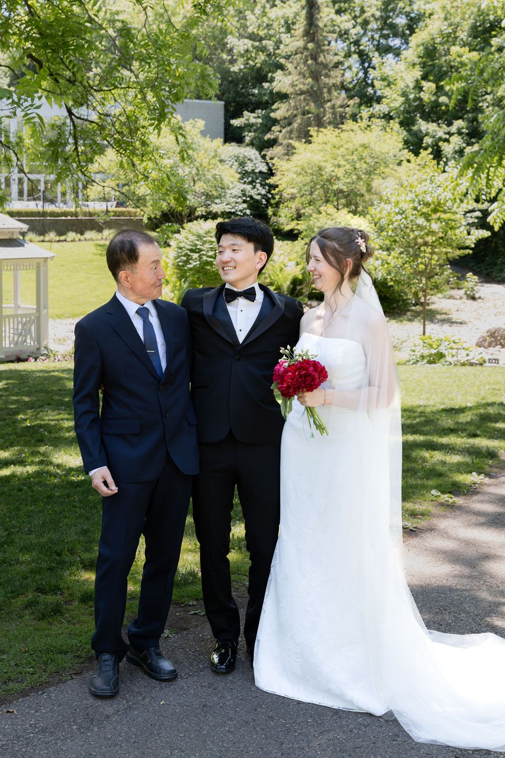 Bride and groom stand with groom father surrounded by greenery at the civic garden complex in london, ontario.