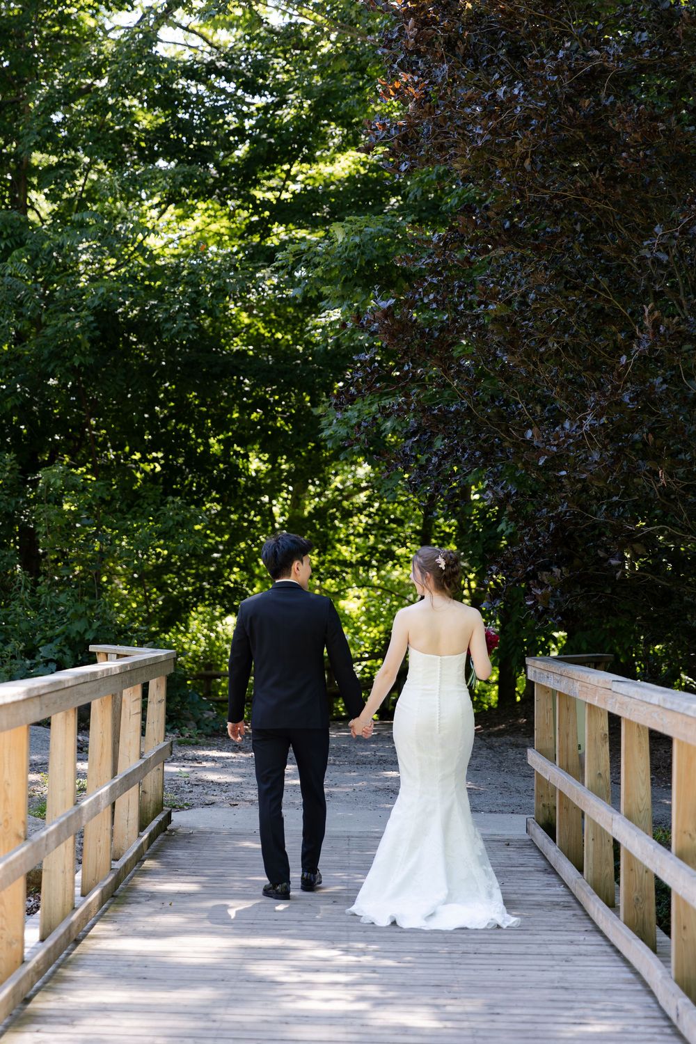 Groom holding brides hand walking across a wooden bridge down a path at the civic garden complex in london, ontario.