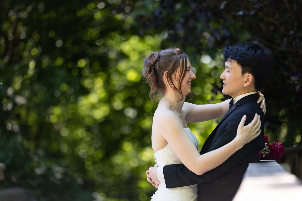 Bride & groom embrace, smiling, surrounded by lush green trees on their wedding day at the civic garden complex in london, ontario.