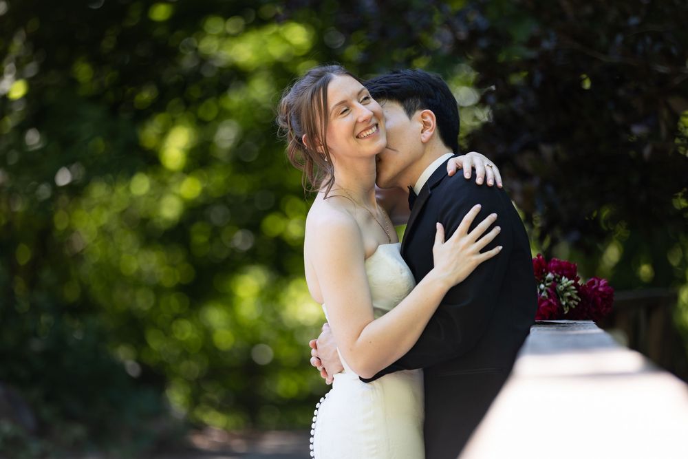 Bride smiles joyfully as a groom in a black suit kisses her cheek outdoors on a trail at the civic garden complex in london, ontario.
