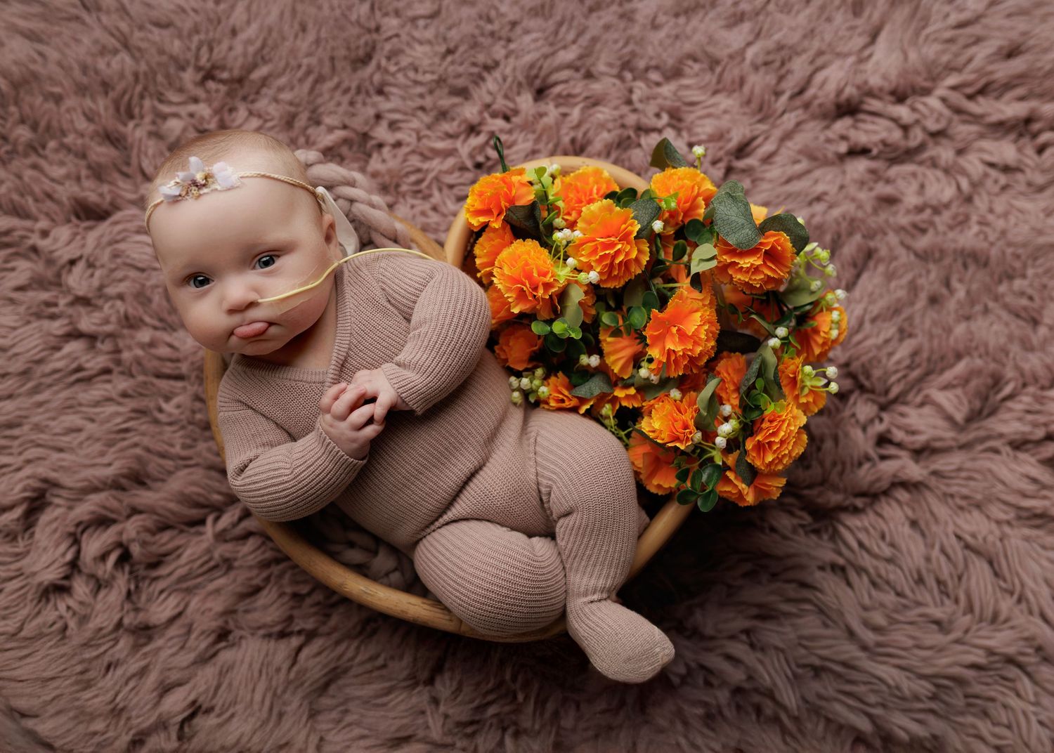 A newborn baby in a tan outfit rests in a wooden bowl beside orange roses on a fluffy mauve rug.