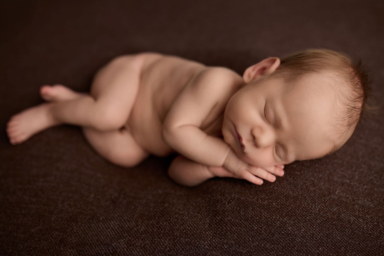 Peaceful newborn baby sleeping curled up on a brown textured blanket in a professional studio portrait.