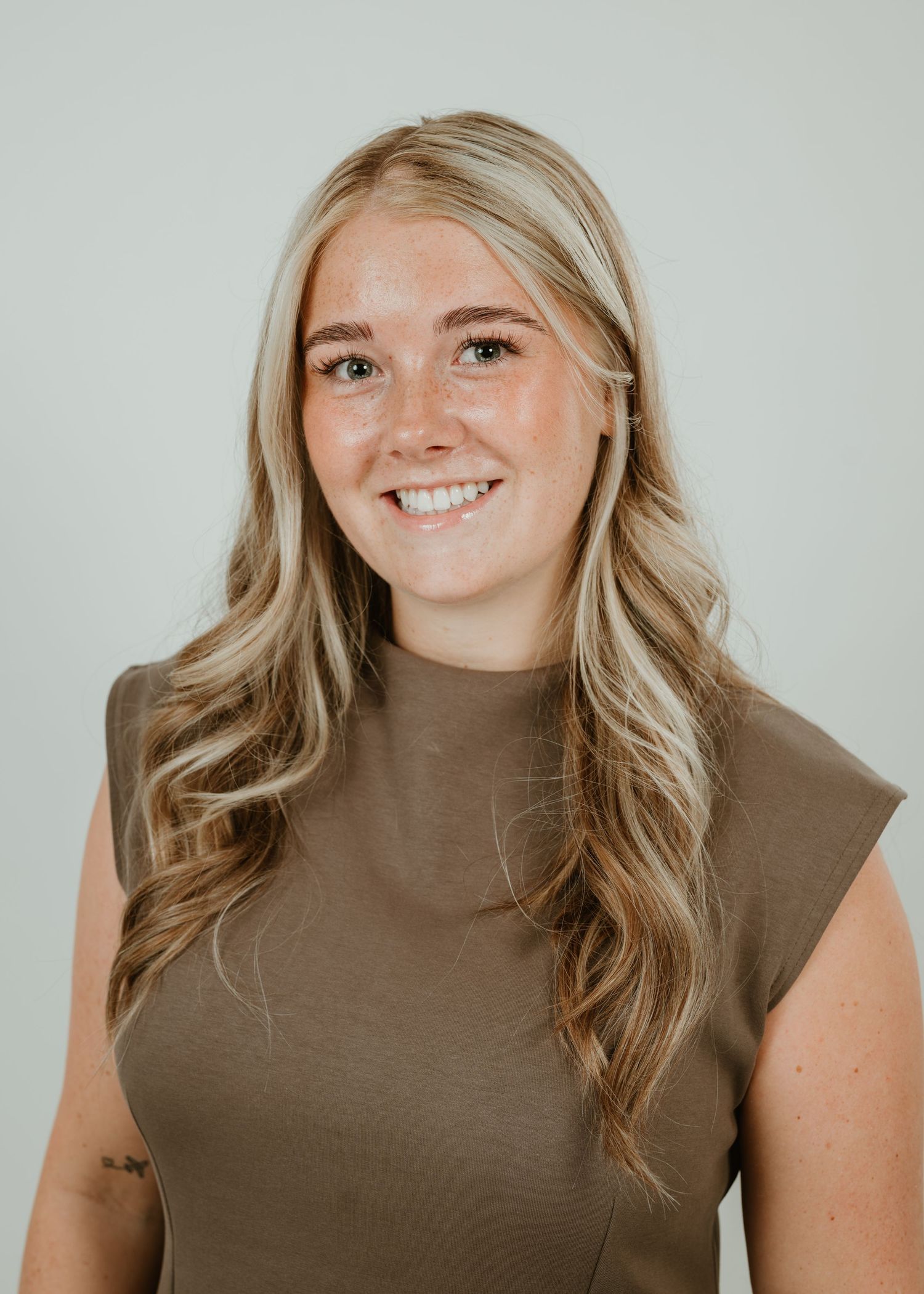 Smiling young woman with long blonde hair wearing a brown sleeveless top against a light gray background.