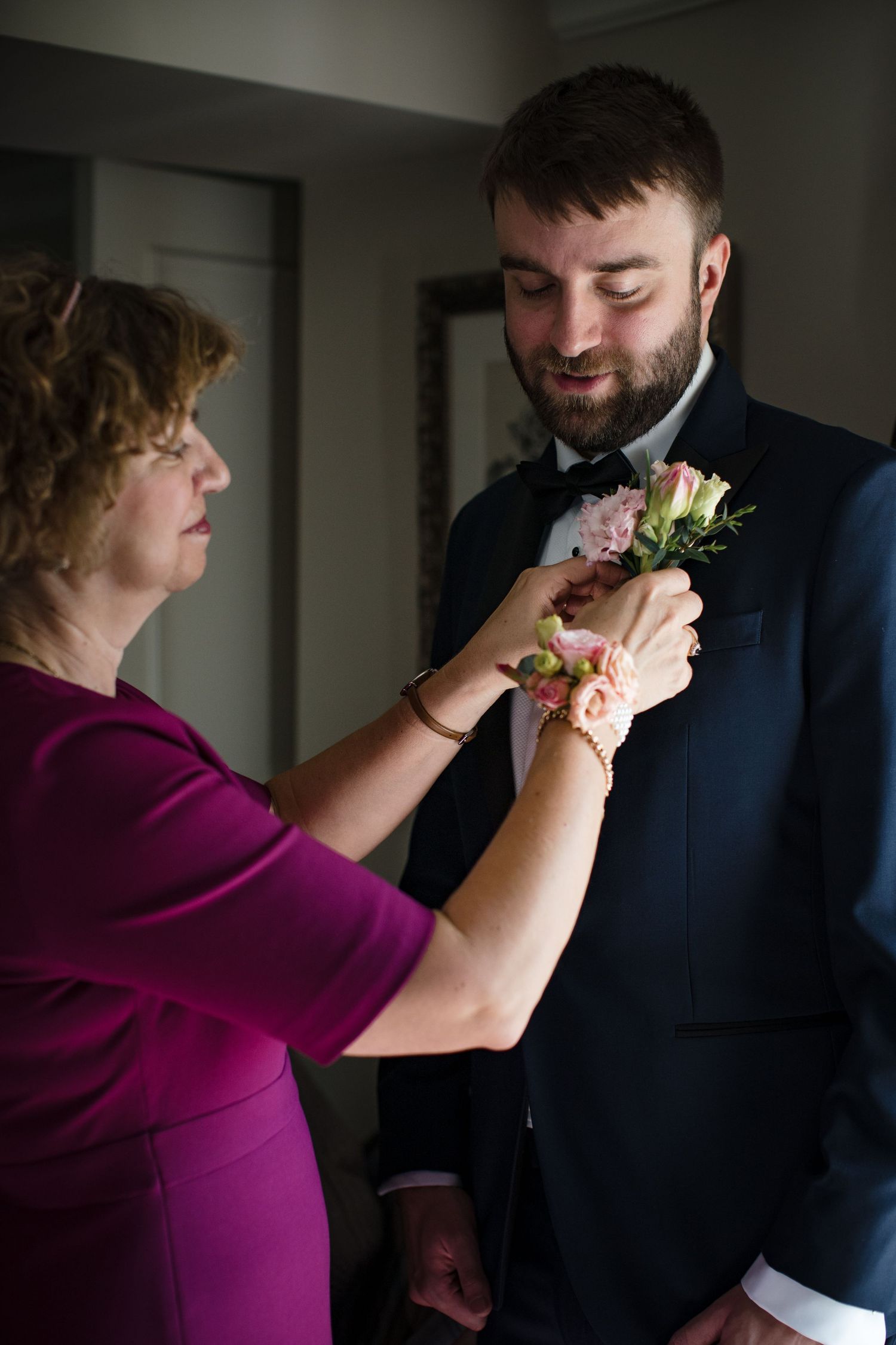 A woman in magenta pins a pink rose boutonniere on a bearded groom in a navy suit before his wedding.