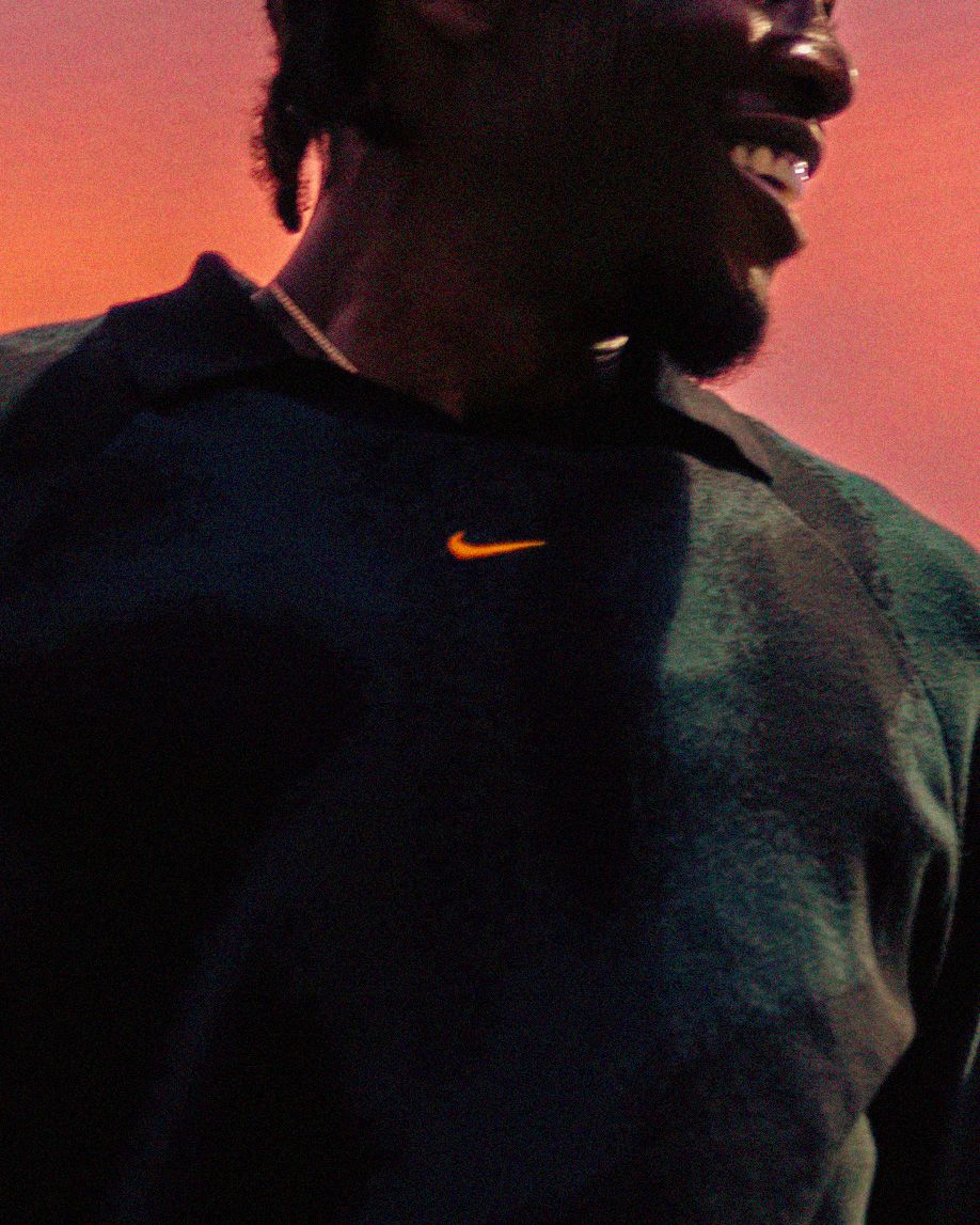 Man smiling wearing Nike dark sweatshirt with orange swoosh at sunset.