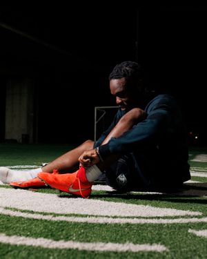 A soccer player sits on a turf field at night, adjusting his bright orange Nike cleats under stadium lights.