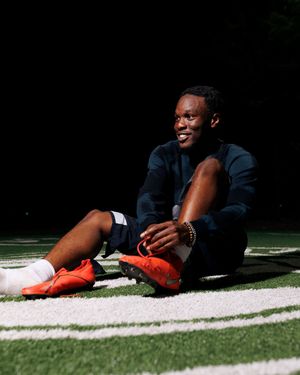 Young athlete in navy outfit sits smiling on turf field wearing orange Nike cleats and white socks.