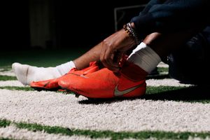 Person lacing up bright orange Nike cleats on artificial turf, wearing white socks and beaded bracelet.