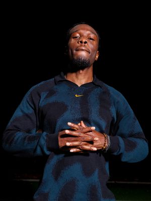 A man in a Nike blue camo sweatshirt stands with hands clasped to chest, eyes closed, against dark background.