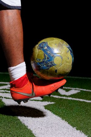 Close-up of a soccer player in orange Nike cleats balancing a worn yellow and blue soccer ball on their foot.