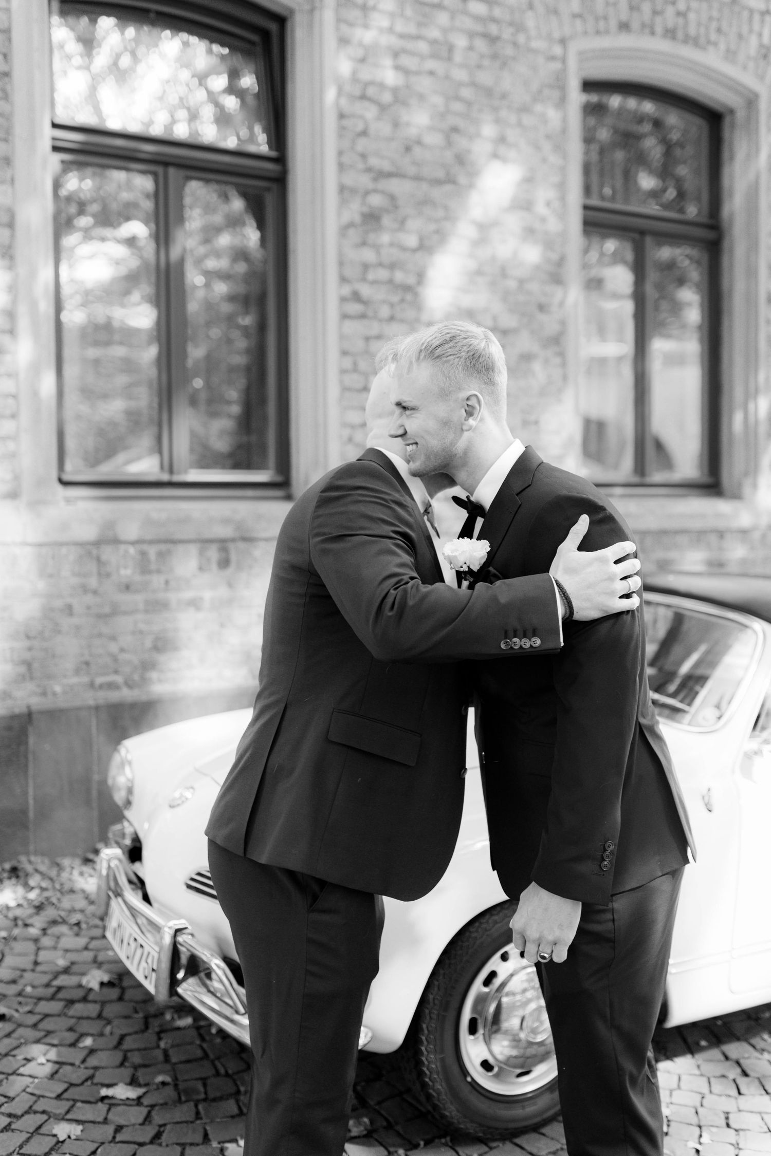 Two grooms in black suits share a warm embrace beside a vintage white car outside a brick building.