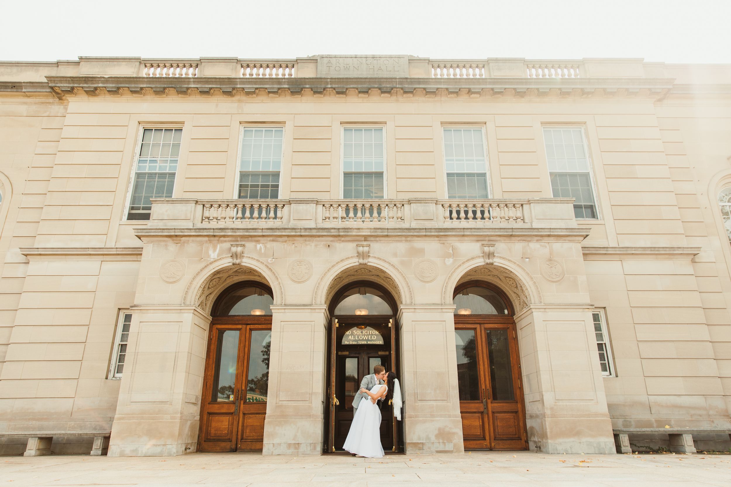 A wedding couple standing in front of a grand limestone building with arched doorways.