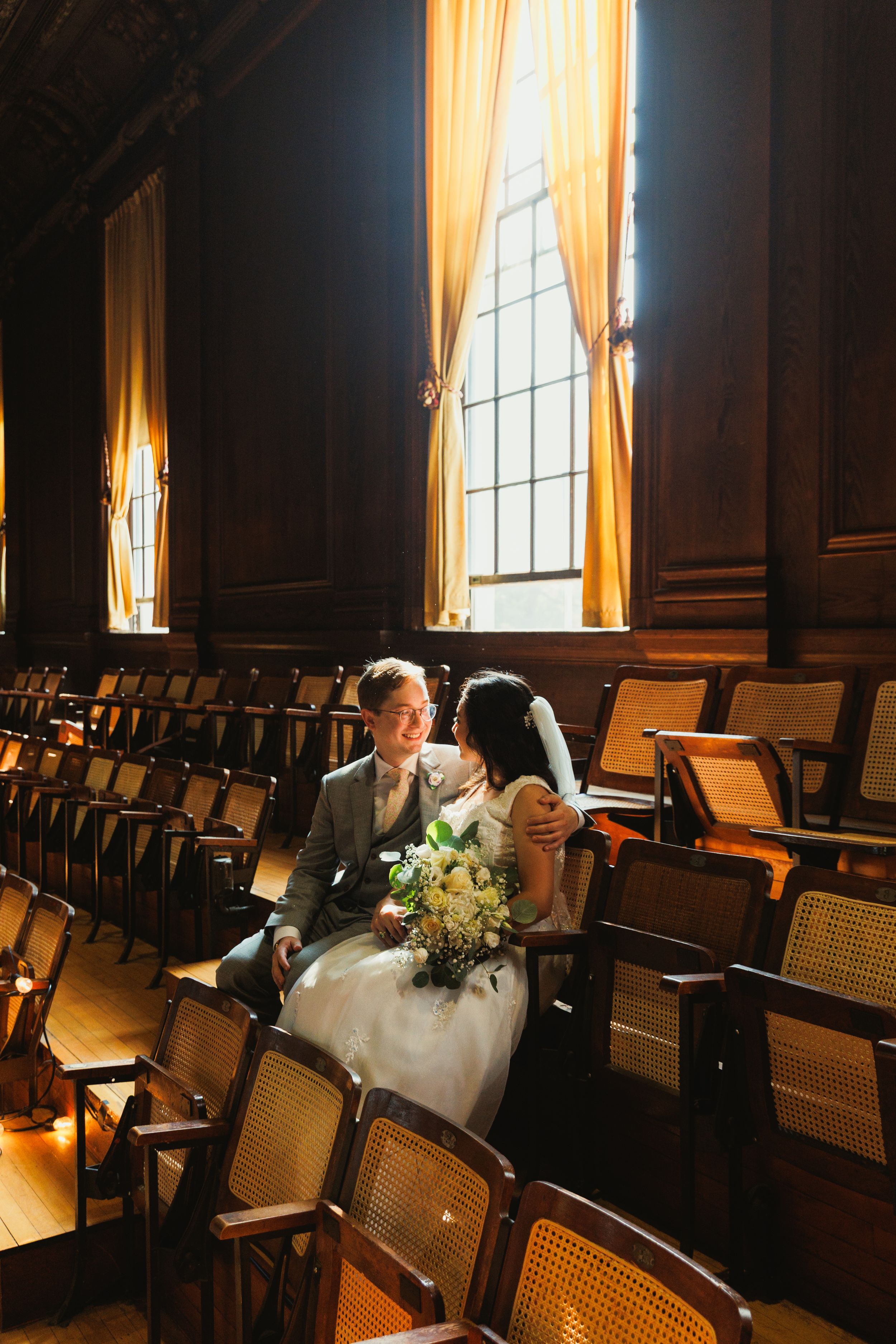 A couple seated in vintage theater chairs with sunlight streaming through tall windows.