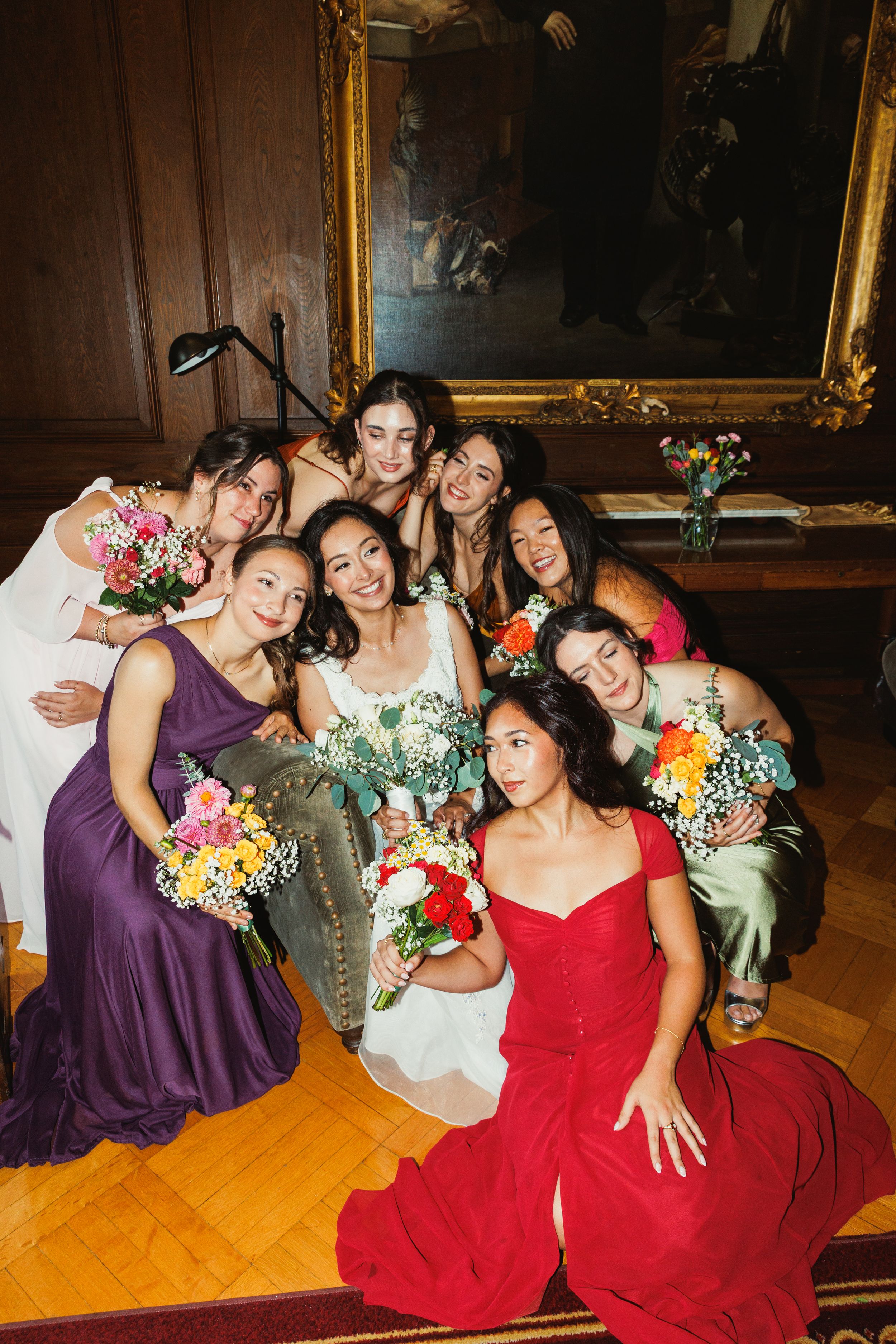A bridal party in purple and red dresses poses together with colorful bouquets in an elegant room with wood paneling.