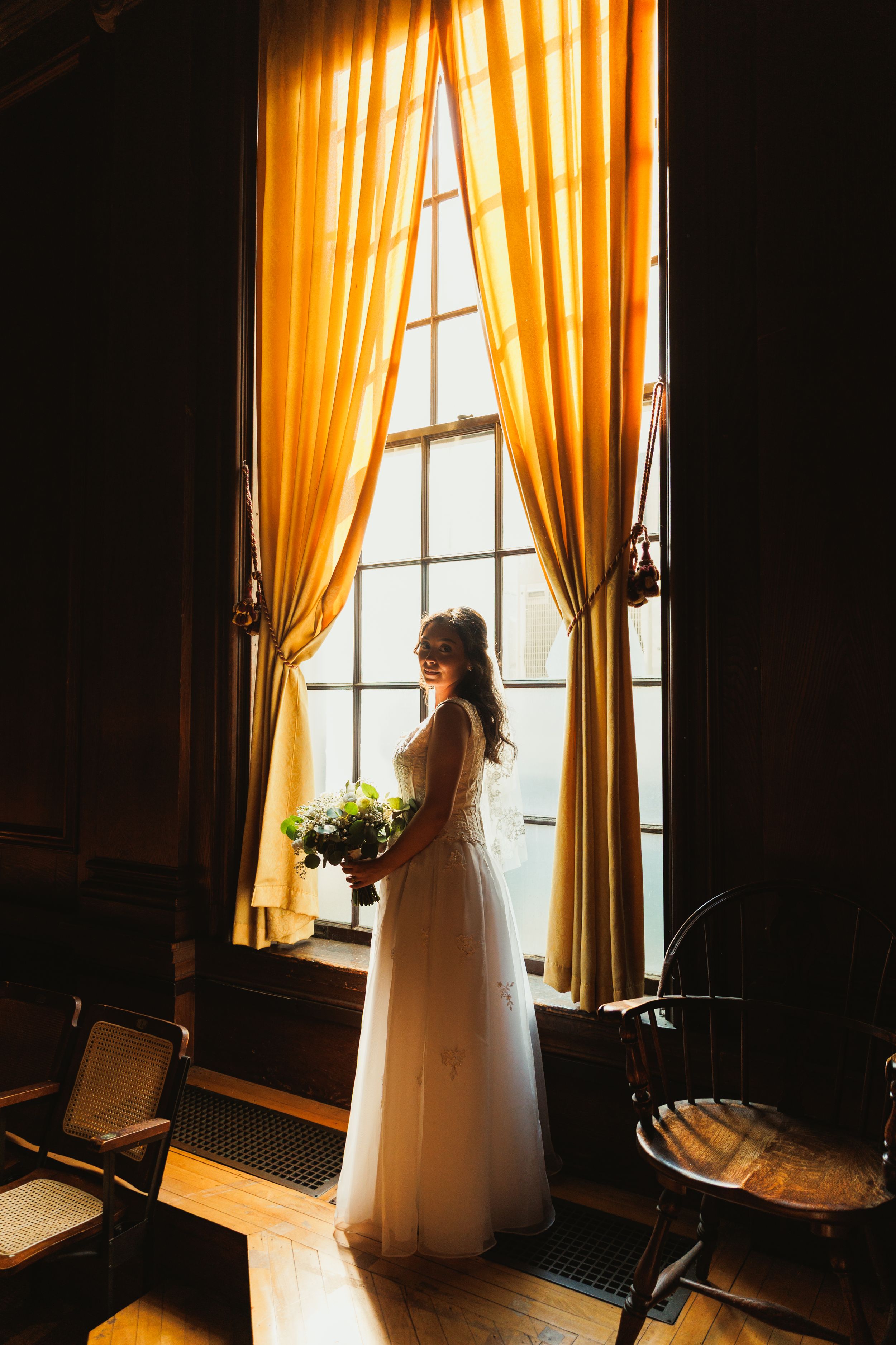 A silhouette of a bride in a flowing gown stands by a window with golden curtains holding a bouquet.