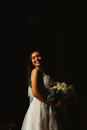 A bride in a white lace dress poses with her bouquet against a dark background.