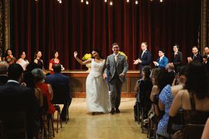 Newlyweds walk down the aisle while wedding guests celebrate in an ornate venue.