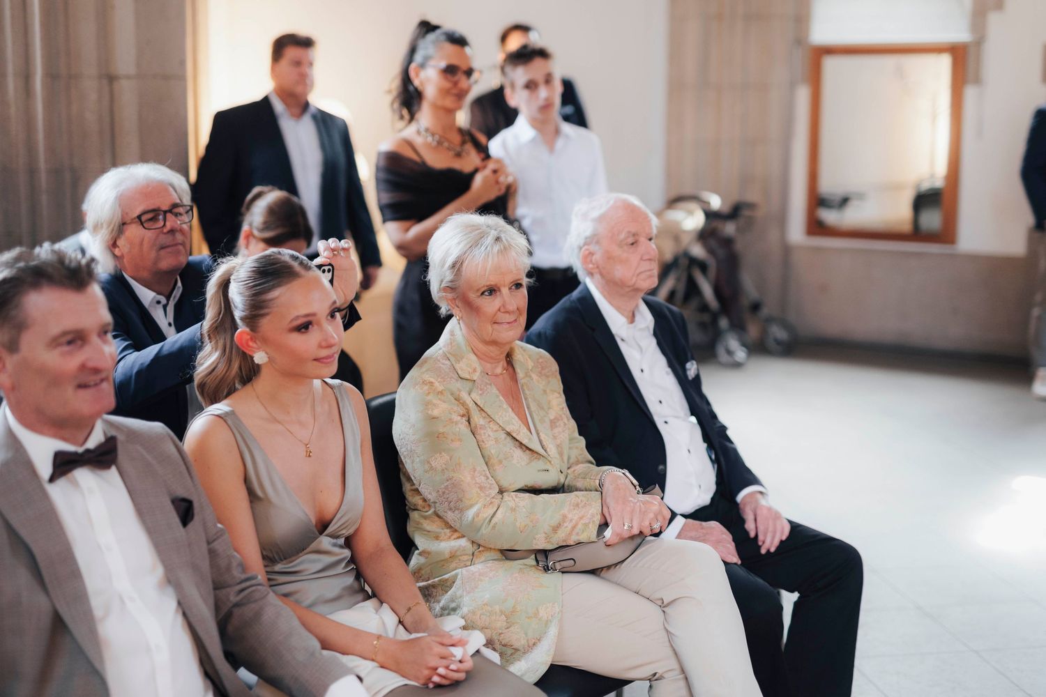 Elegantly dressed wedding guests seated in rows, smiling during a ceremony in a bright, modern venue.