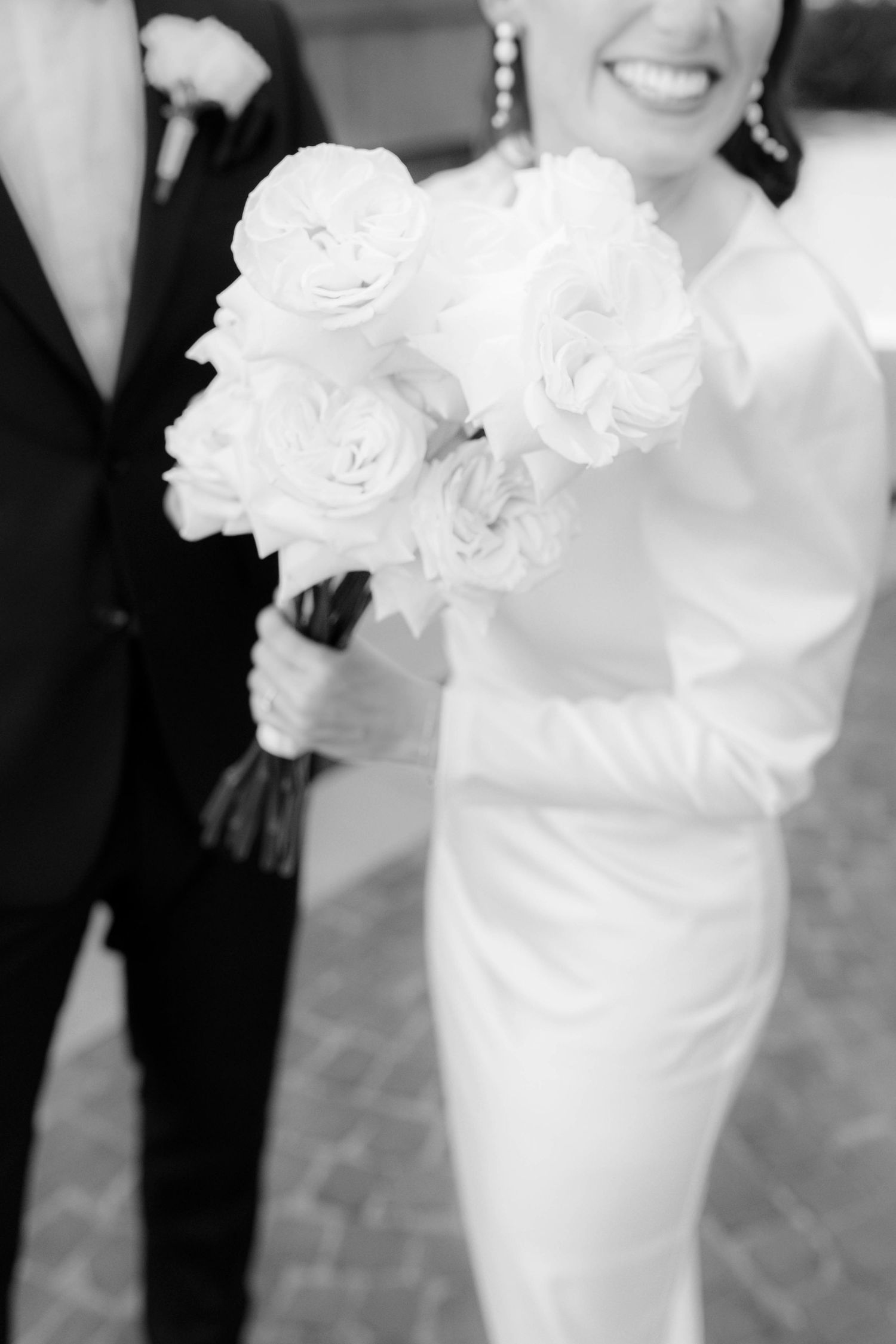 Black and white photo of a smiling bride in white gown holding a bouquet of white roses beside a groom in dark suit.