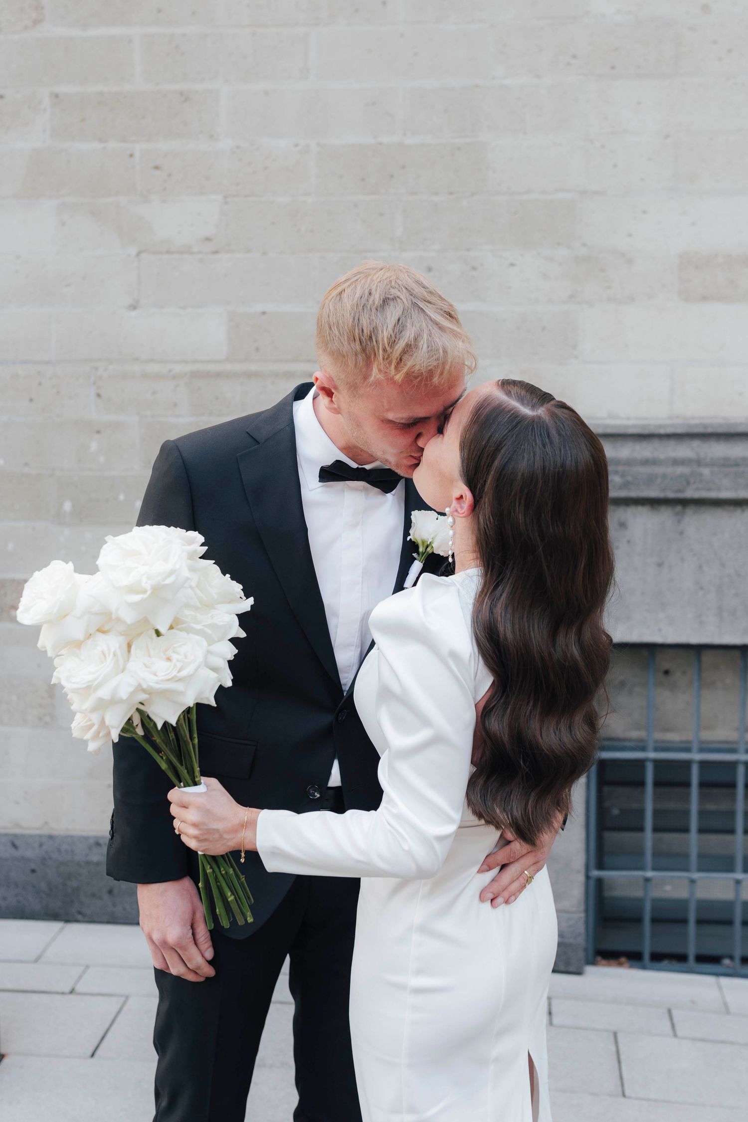 Bride and groom sharing a kiss, she holds white peony bouquet, he wears black tuxedo, outdoor stone building backdrop.
