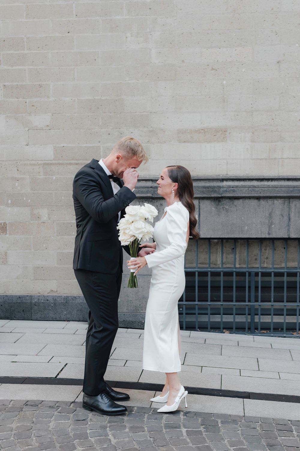 Emotional groom wiping tears as bride in white dress holds bouquet of white roses during wedding first look.
