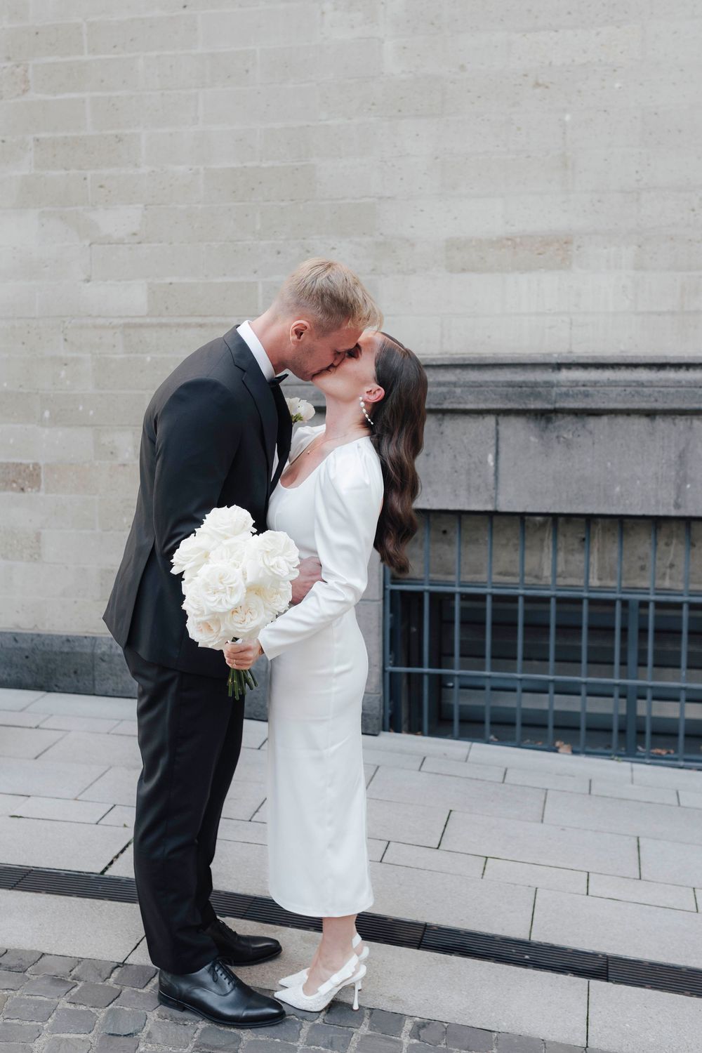Bride and groom sharing a kiss outdoors, bride in white midi dress holding white peony bouquet, groom in black tuxedo.