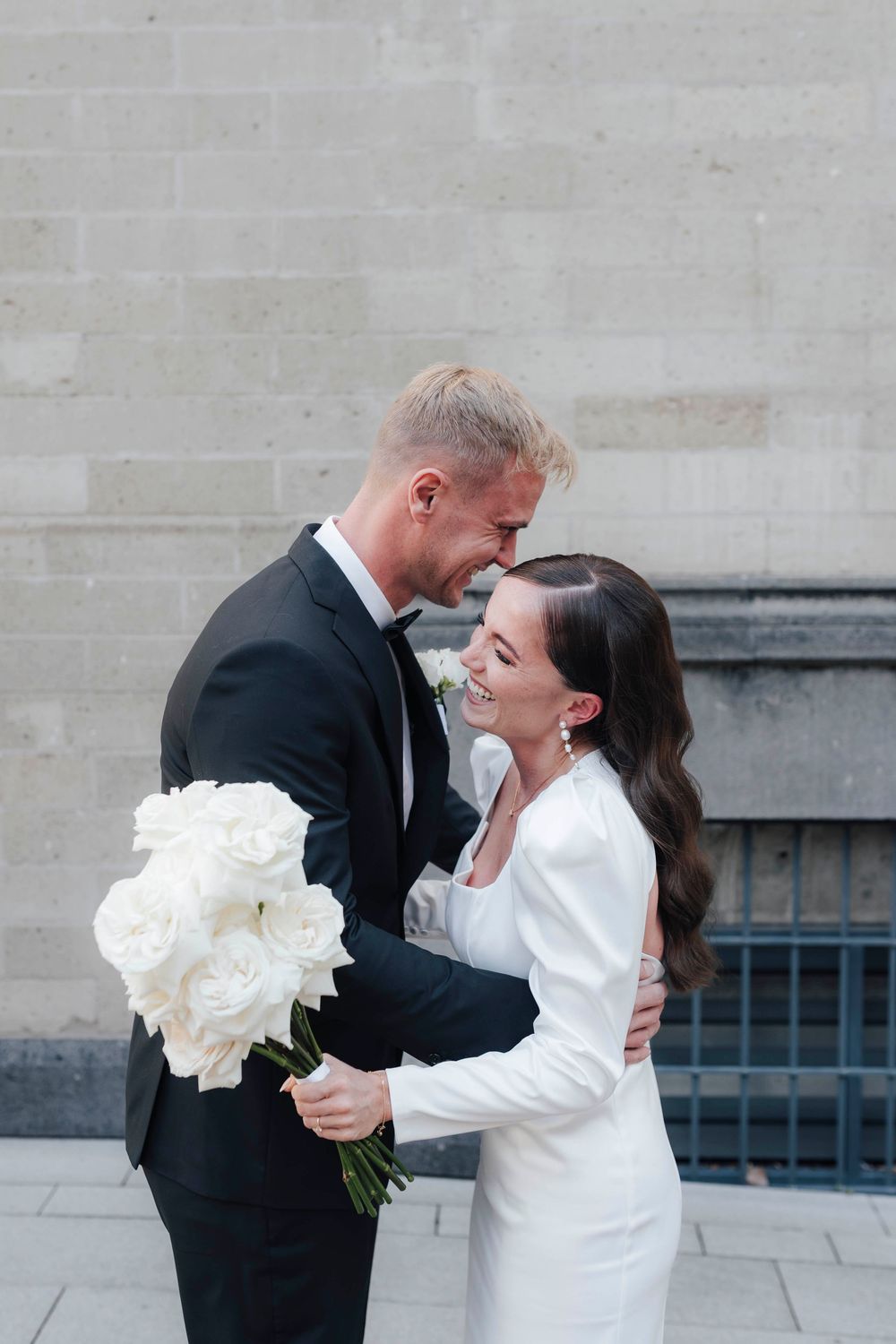 Laughing bride and groom embracing outdoors, bride holding white rose bouquet in elegant wedding attire.