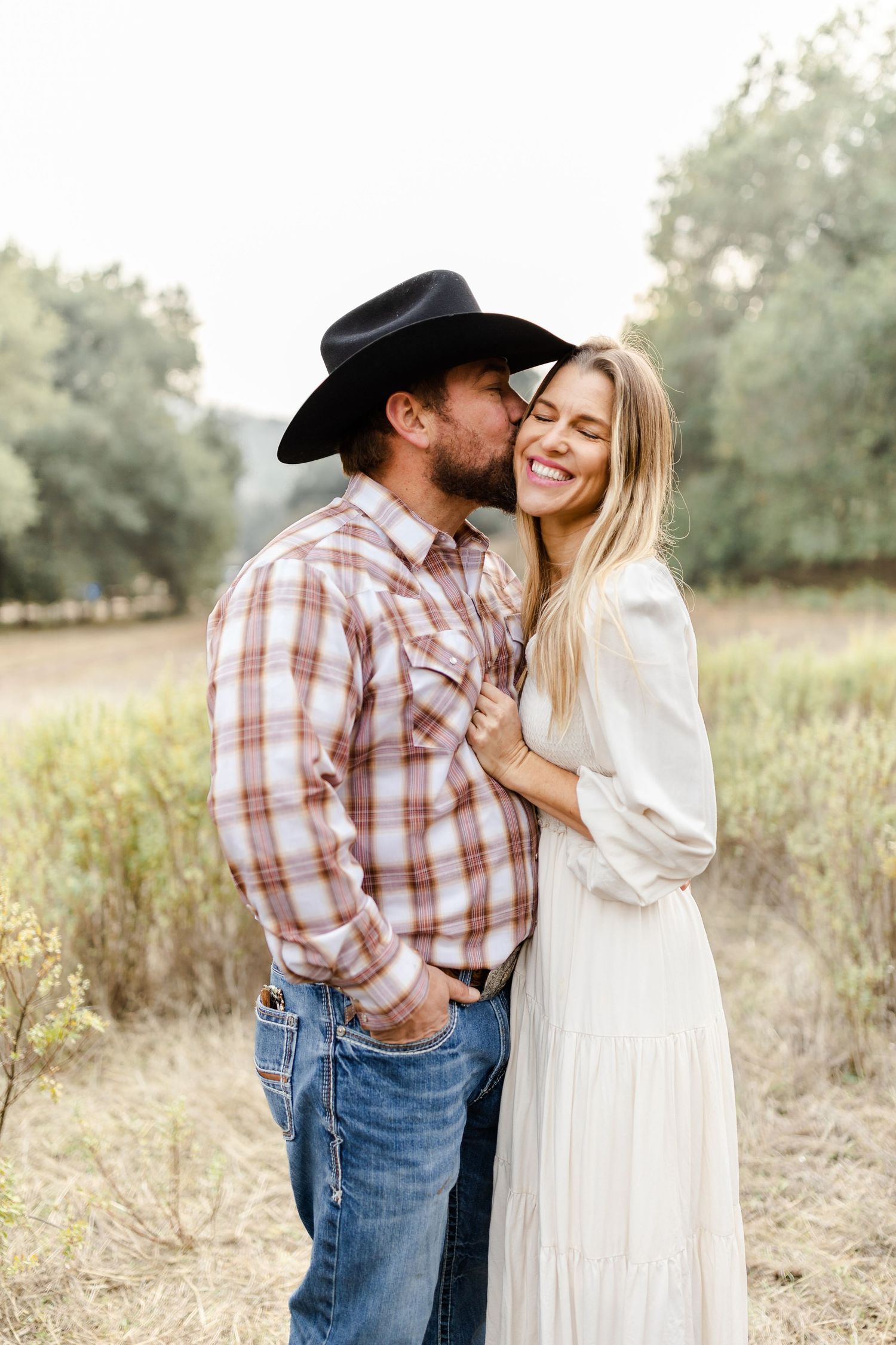 A couple poses outdoors, man in cowboy hat and plaid shirt kisses woman in white dress who smiles joyfully.