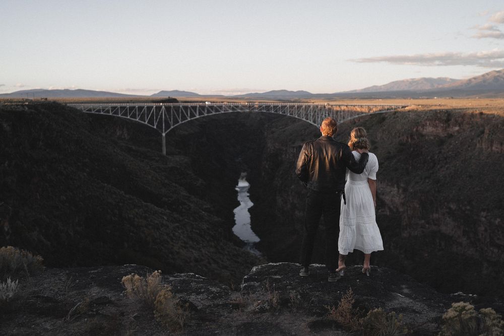 Bride and groom embracing on rocky cliff overlooking Rio Grande Gorge Bridge at golden hour sunset