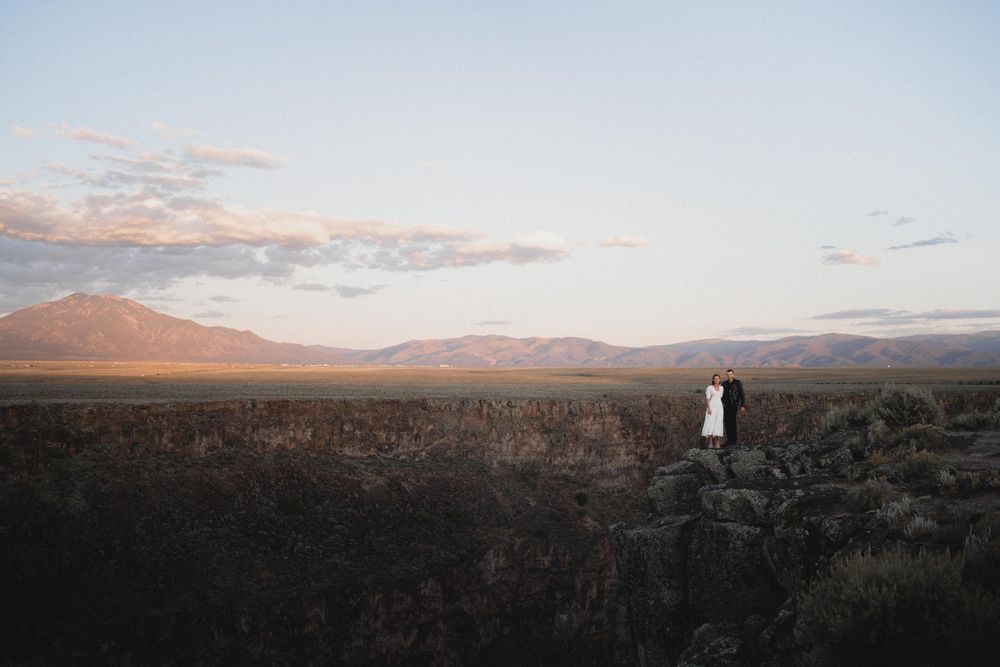 Couple standing on rocky desert landscape at sunset with vast mountain valley and dramatic sky behind them at Taos Gorge
