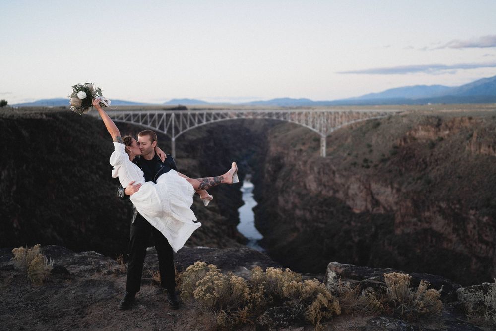 Groom dipping bride on canyon cliff edge with Rio Grande Gorge Bridge and river visible below.