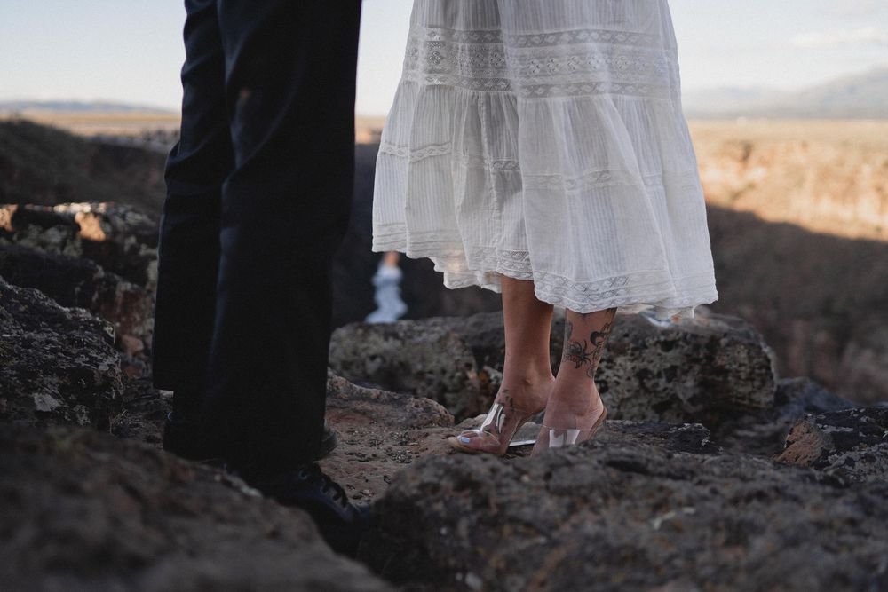 Close-up of bride in white lace dress and clear heels standing on rocky terrain with groom in dark suit.