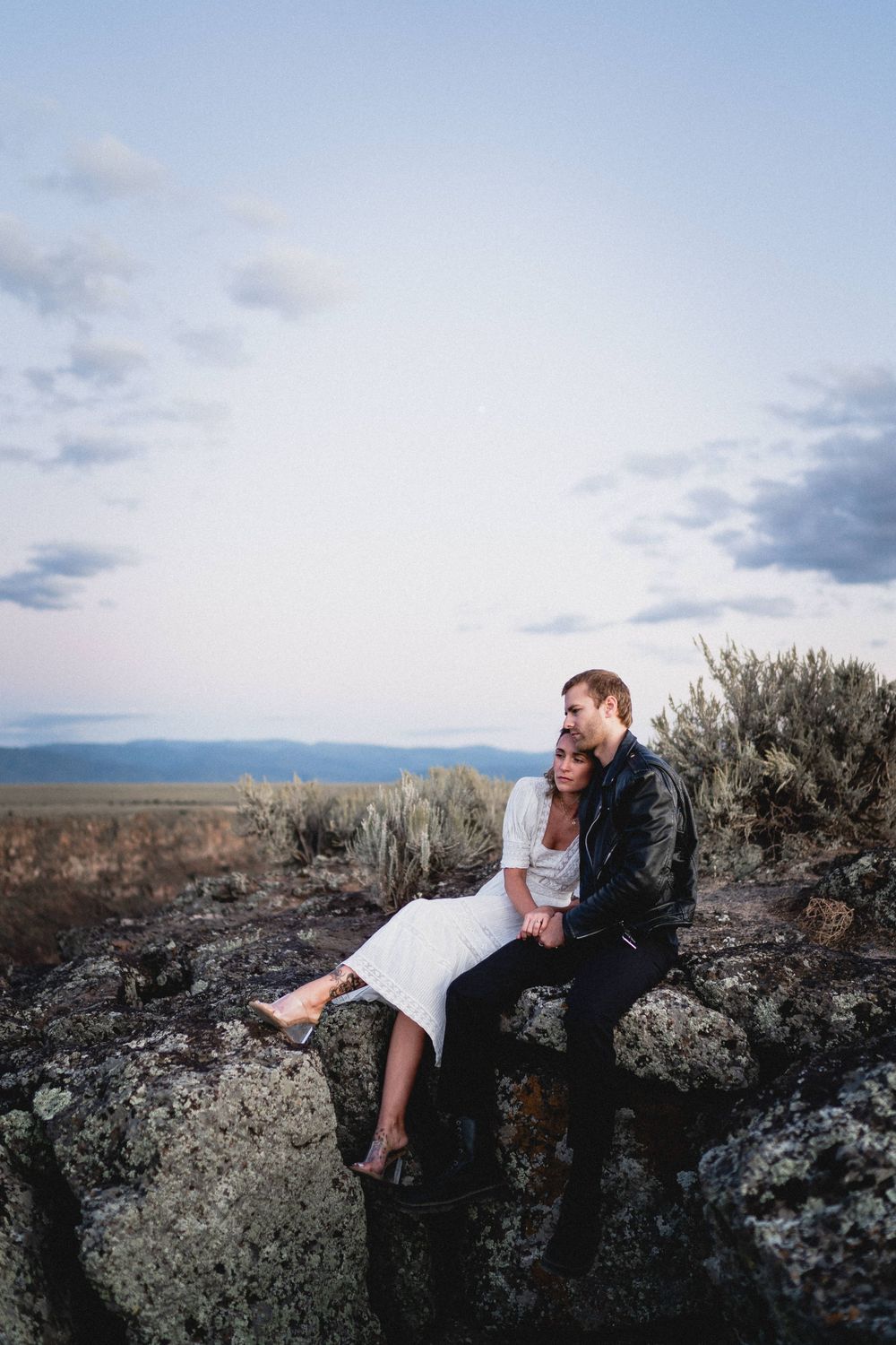 Couple sitting closely together on rocky outcrop surrounded by sagebrush under a soft evening sky at Rio Grande Gorge in Taos New Mexico