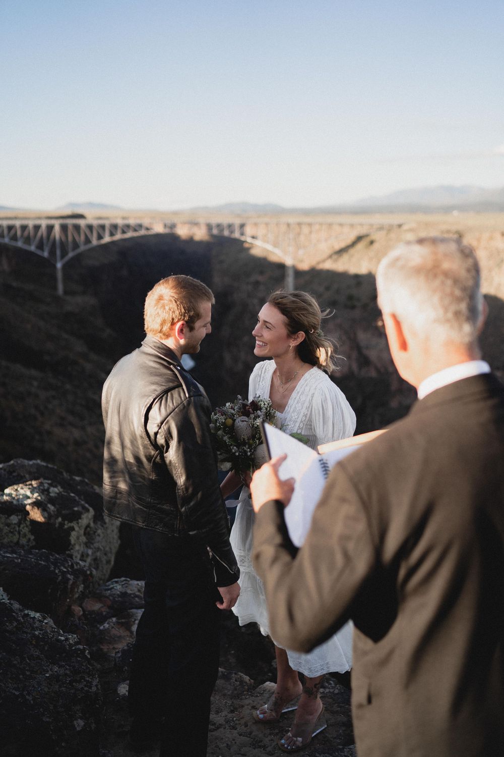 Smiling bride and groom exchanging vows on rocky cliff with iconic Rio Grande Gorge Bridge behind them.