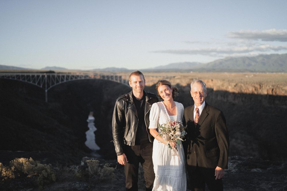 Wedding party of three posing outdoors with Rio Grande Gorge Bridge and mountains in background at sunset in Taos