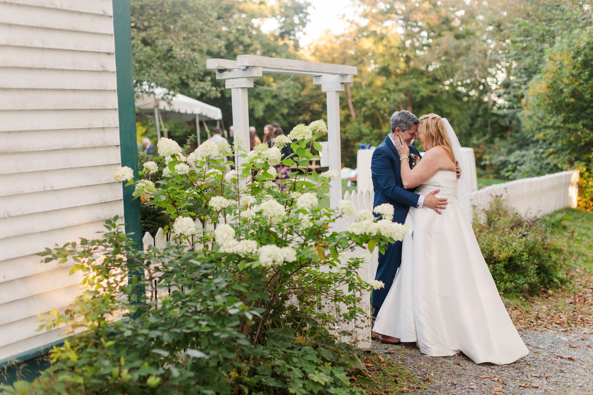 Bride in a white gown and groom in a dark suit embrace beside blooming white hydrangeas near a garden pergola at an outdoor wedding.
