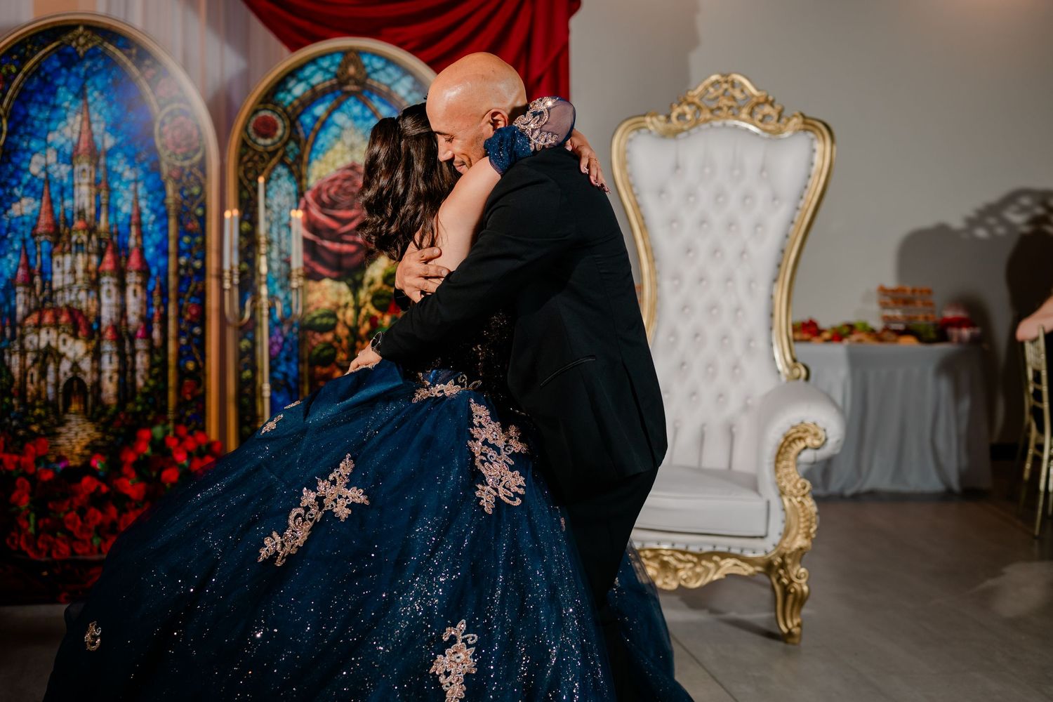Father embraces a young woman in a navy blue embroidered quinceañera gown beside a white throne chair and stained glass panels.