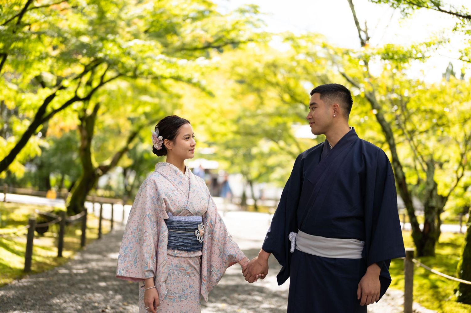 Couple in traditional Japanese kimono holding hands while walking through a serene tree-lined garden path.