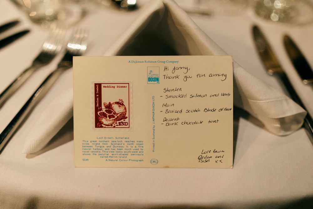A handwritten postcard with a vintage stamp displayed on a white tablecloth at a formal dinner setting.