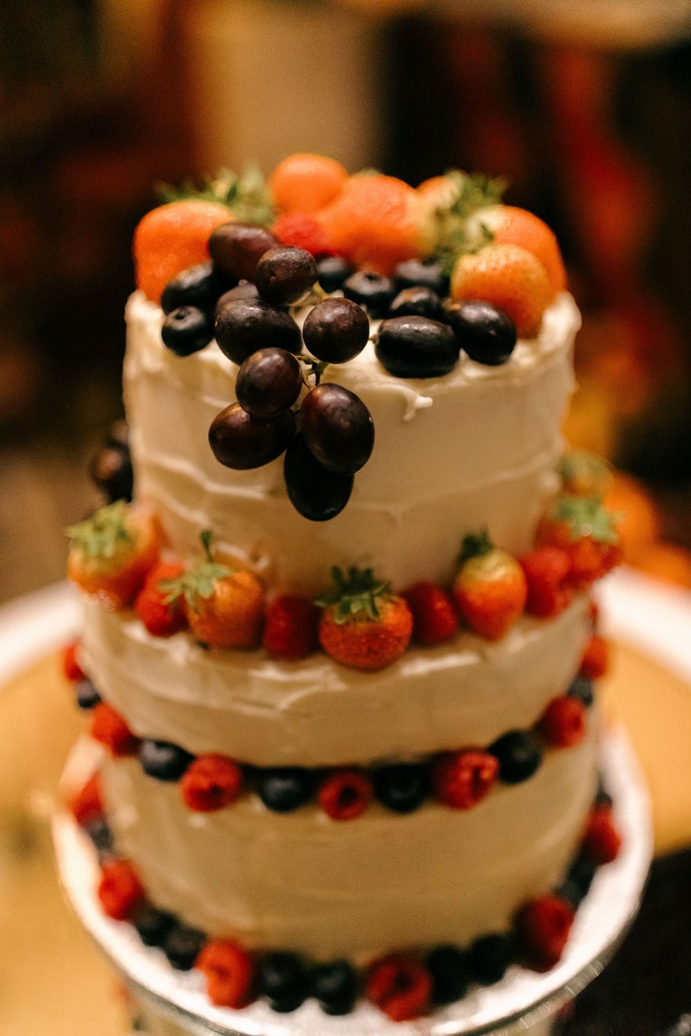 Three-tier white frosted wedding cake decorated with fresh strawberries, blueberries, raspberries, and grapes.