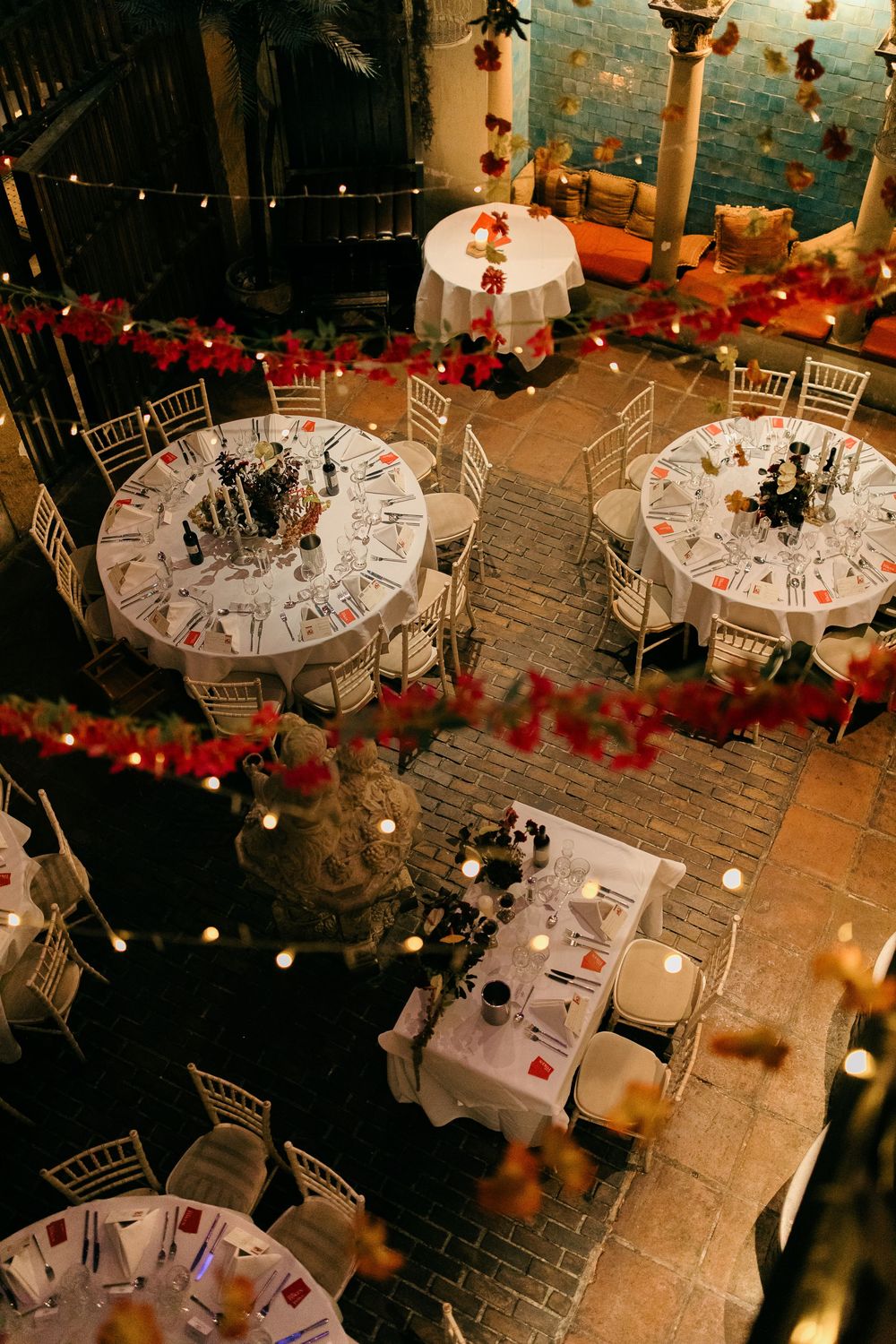 Elegant wedding reception venue with round tables, white linens, chiavari chairs, red floral garlands, and warm candlelight.