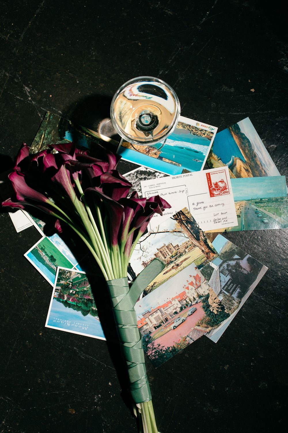 Dark purple calla lily bouquet with vintage postcards and champagne glass on black marble surface.