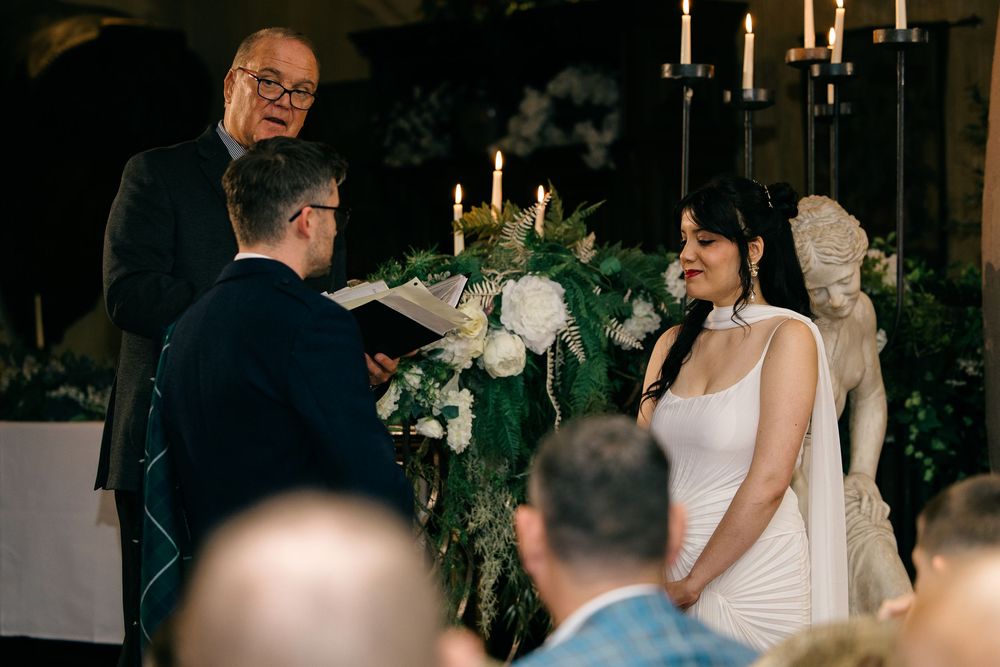 Couple exchanging wedding vows at candlelit altar with white floral arrangements, officiant reading ceremony script.