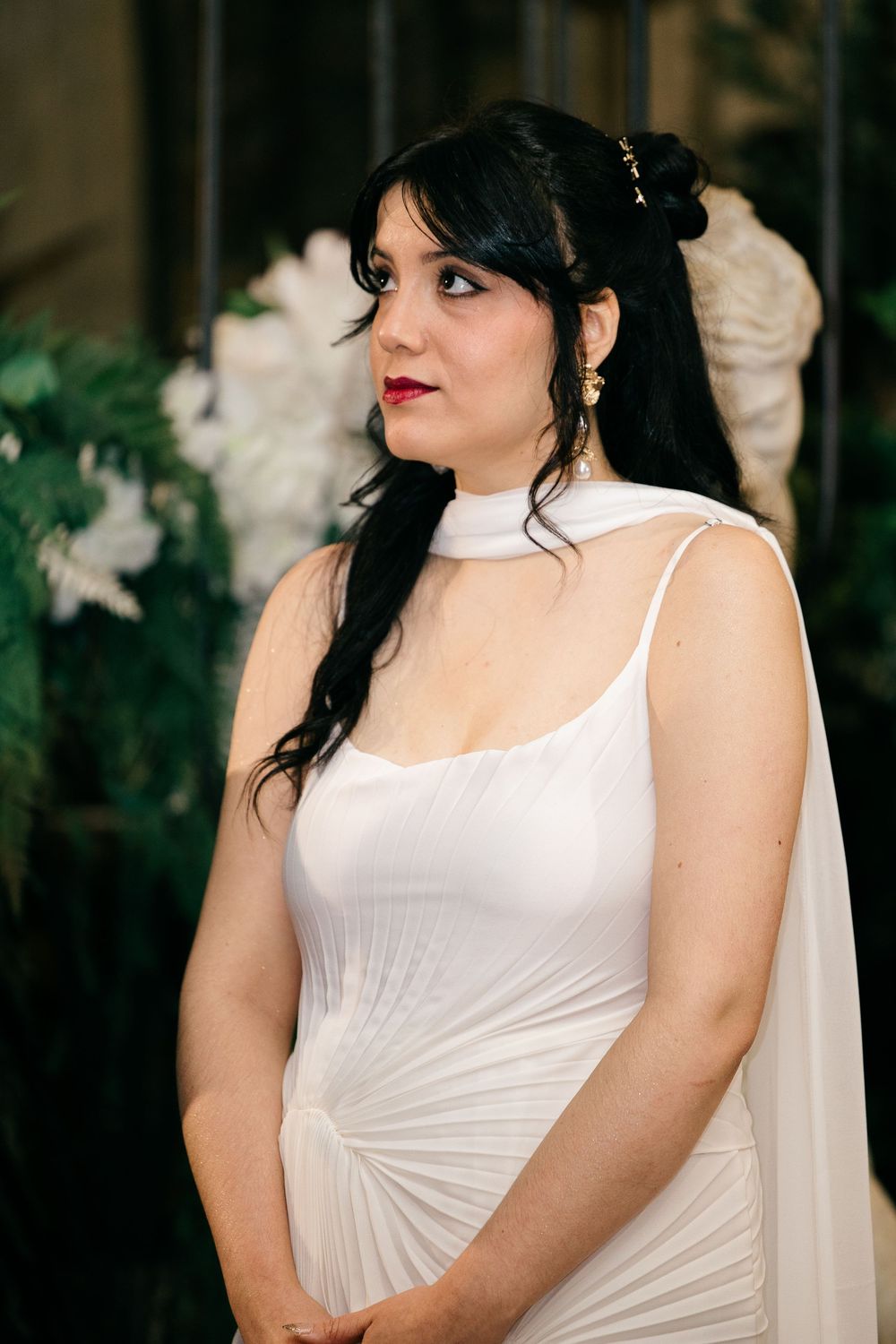 Bride in white fitted gown with cape detail and gold hair accessories against lush green backdrop.