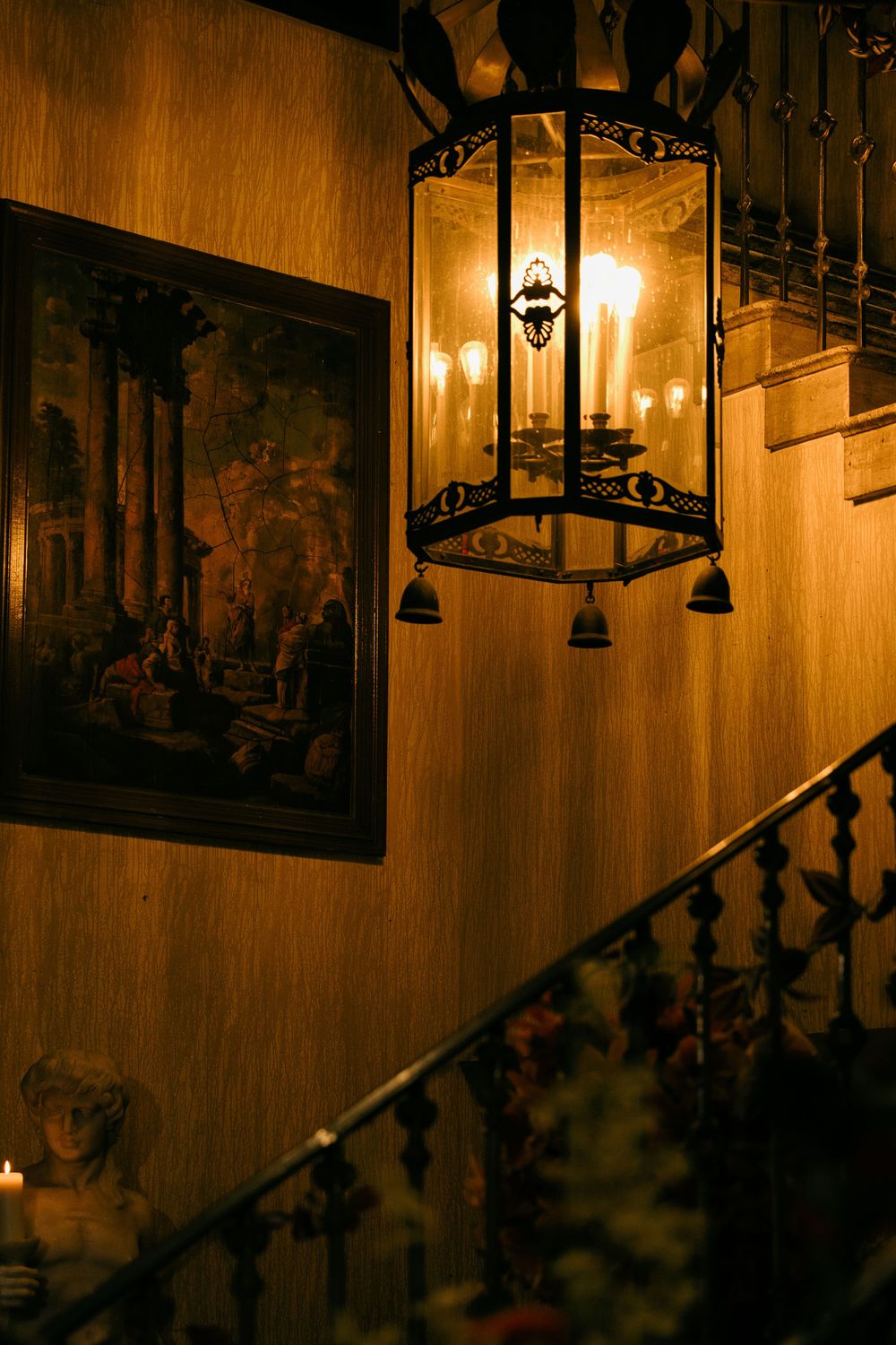 Ornate black iron lantern with candles illuminating a grand staircase beside a classical painting in warm golden light.