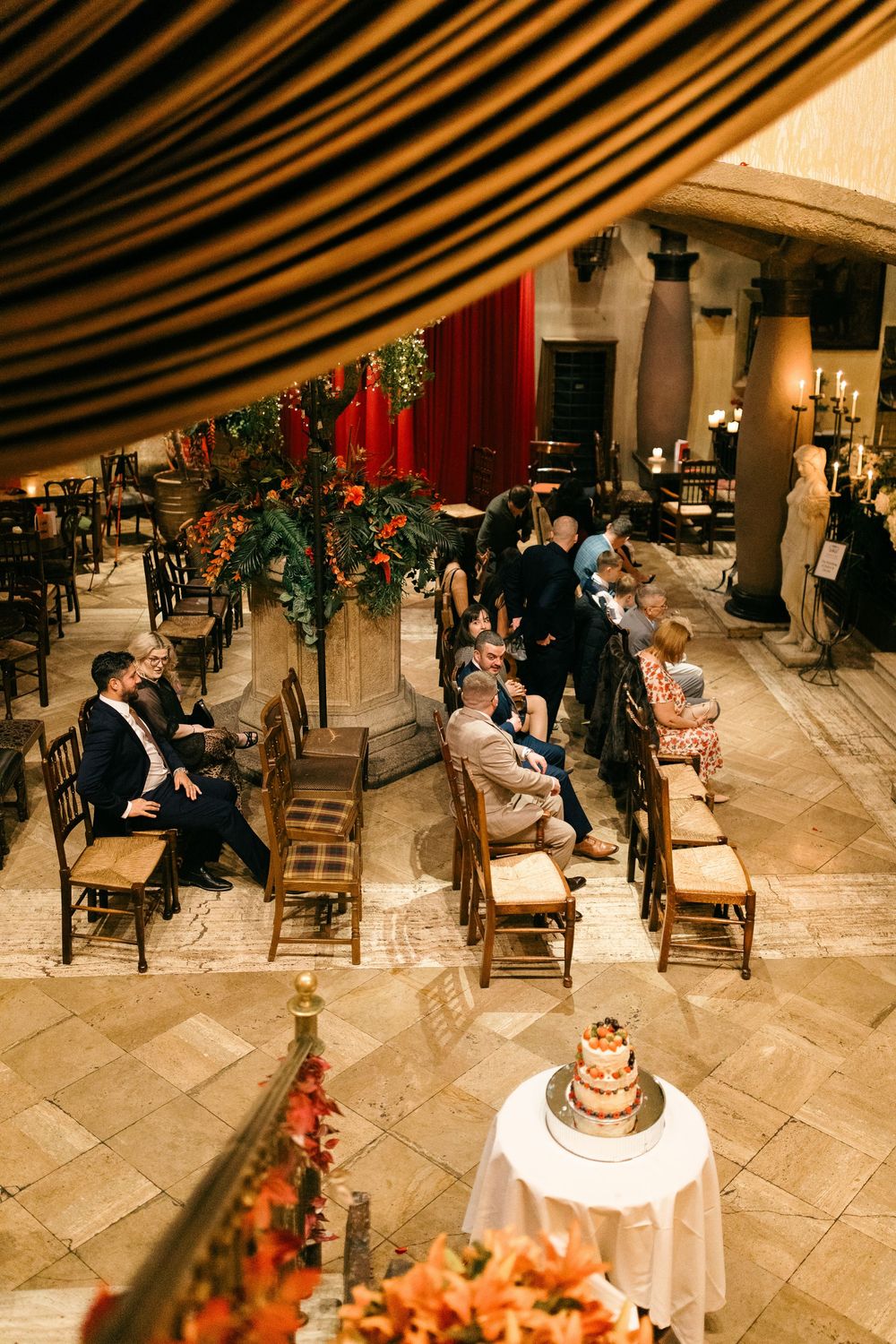 Aerial view of elegant wedding venue with guests seated in wooden chairs, tiered cake, and floral arrangements in warm tones.