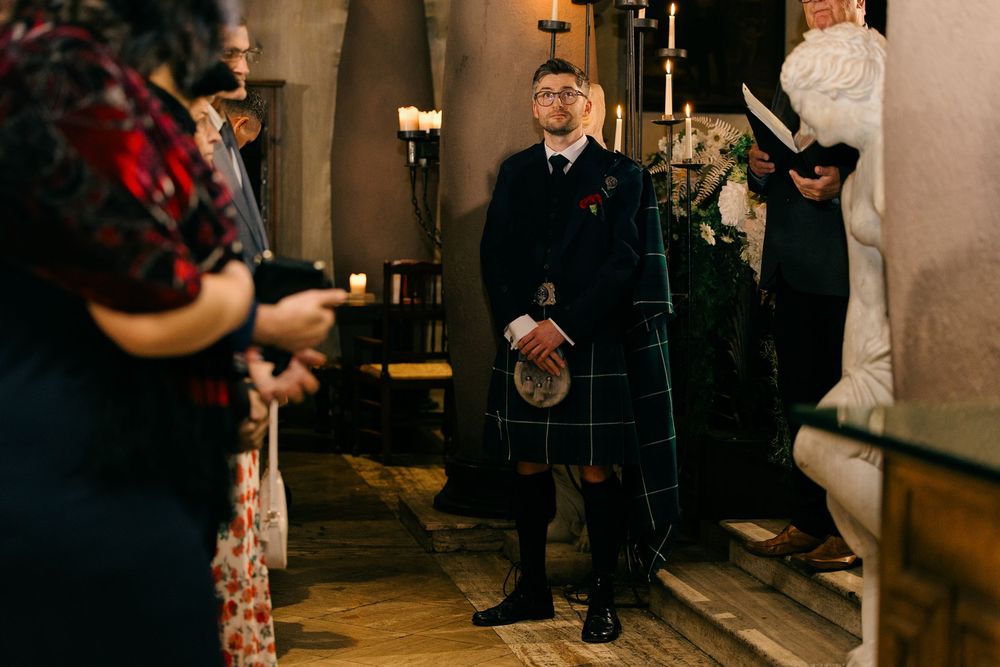 Groom in traditional Scottish kilt stands at wedding ceremony aisle in historic stone church.