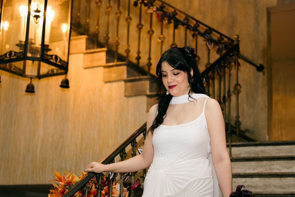 Bride in white gown poses gracefully on ornate staircase with chandelier in elegant venue.