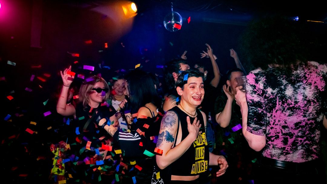 People dancing at a neon-lit nightclub party with colorful confetti falling under a disco ball.