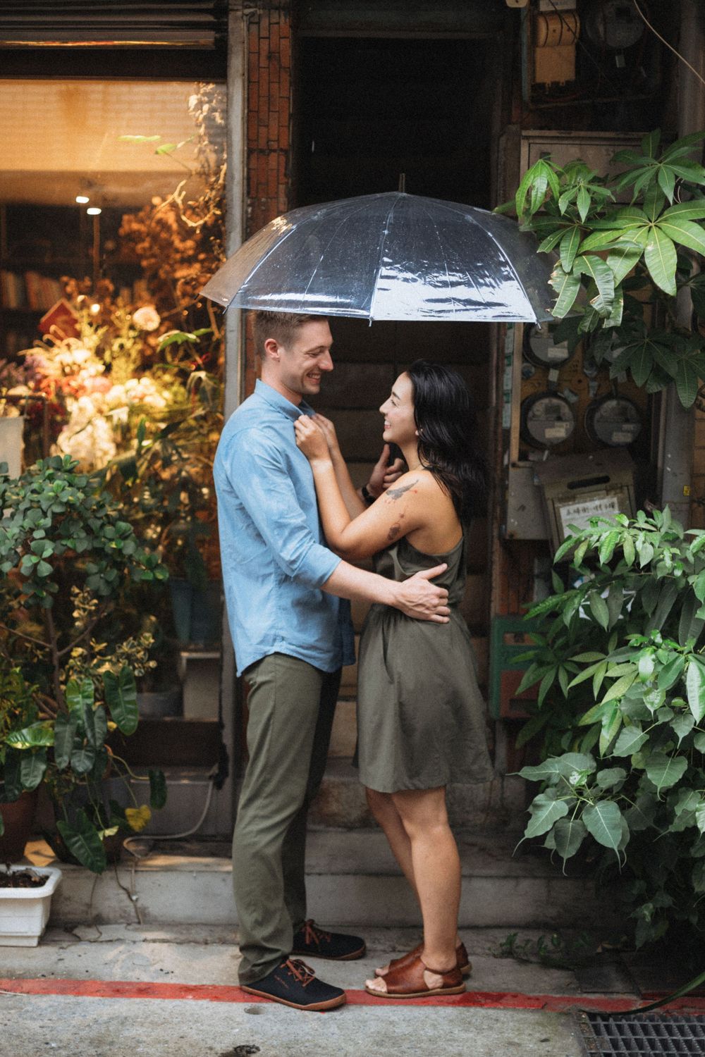 Couple sharing a clear umbrella outside a flower shop, smiling at each other in the rain surrounded by lush green plants.